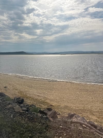 This image depicts a serene natural landscape. The scene is divided into three main sections: the sky, the water, and the shore. The sky occupies the left portion of the image and is filled with a dramatic display of white and gray clouds, suggesting a dynamic and possibly changing weather pattern. The clouds are textured and voluminous, with sunlight breaking through in places, creating a sense of depth and luminosity.
The central part of the image is dominated by a vast body of water, likely the sea given the location context of Port-la-Nouvelle, France. The water's surface is shimmering, reflecting the light from the sky, and gentle waves are visible near the shore. The horizon line is low, emphasizing the expanse of the water.
The right side of the image showcases the shore, which appears to be a sandy beach. The sand has a fine texture and a light, earthy color. Towards the right edge, there are darker, rocky areas interspersed with some dry grass and vegetation, suggesting a natural, undeveloped coastline. The transition from sand to rock and vegetation is abrupt in places.
The image appears to have been taken during the daytime, possibly late afternoon or early morning, given the quality of light and the presence of shadows. The overall mood is tranquil and contemplative, with the natural elements of sky, water, and land creating a harmonious scene. There is no visible text in the image. The lack of people or man-made objects contributes to the feeling of solitude and natural beauty. The context of Port-la-Nouvelle, France, places this scene within a coastal environment known for its beaches and natural landscapes.