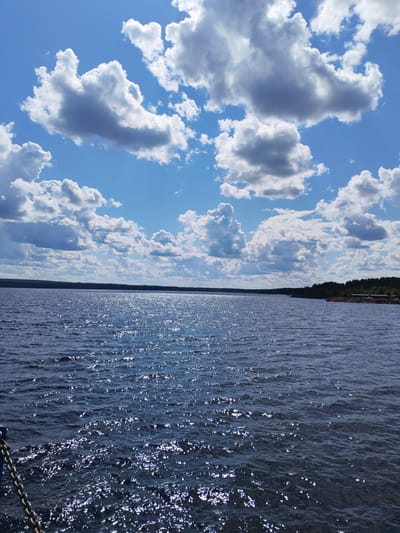 A wide, still lake stretches towards a distant tree-lined horizon under a bright blue sky dotted with fluffy white clouds. Sunlight glints off the dark water, creating a shimmering effect. The scene appears to be a natural landscape, likely in a rural or undeveloped area. There are no people or specific landmarks visible that would indicate the city of Kvartsitny, Russia.