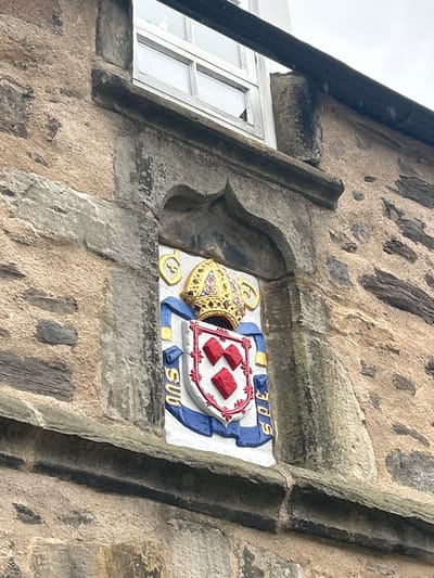 The image shows a heraldic panel set into a stone building, possibly in Aberdeen City, United Kingdom. The panel features a coat of arms with a decorated helmet crest to the left and a shield with a white field bearing three red lozenges within a red border. A blue ribbon-like device surrounds the shield with the letters "SPE" above and "SUB" below. The surrounding building is made of rough, weathered stone, and a window is visible to the left of the panel. The architectural style and materials suggest a historical building. The weather appears overcast.