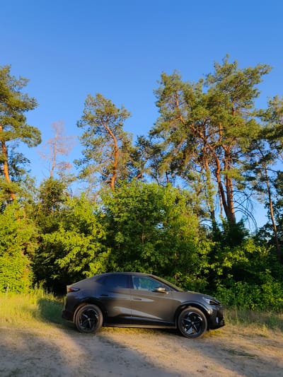 A dark grey electric SUV is parked on a dirt road next to a lush green forest under a clear blue sky. The car, a BYD Yuan, is angled towards the right of the frame, showcasing its sleek design and black alloy wheels. The forest is dense with a mix of pine trees and deciduous trees, with some of the pines appearing tall and slender with reddish-brown bark. The ground is a mixture of dirt and dry grass, with dappled sunlight casting shadows from the trees. The time of day appears to be late afternoon or early evening, indicated by the warm, golden light illuminating the scene. The weather is clear and sunny. The location is Shyroke, Ukraine. No people are visible in the image. No text is visible.