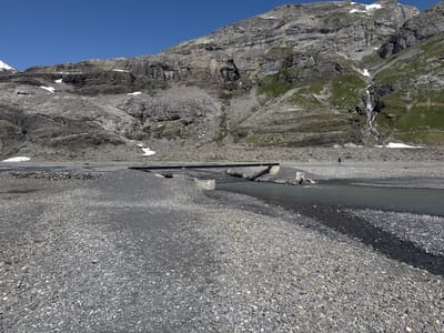 A wide-angle view shows a mountainous landscape in Leukerbad, Switzerland, under a clear blue sky. In the foreground, a wide, grey, gravel-covered riverbed stretches across the frame, with a narrow, dark stream of water flowing through it. A simple, dark-railed bridge crosses the stream further into the background. Beyond the riverbed, a steep, rocky mountain face rises dramatically, showing layers of grey rock and patches of green grass and snow. A thin waterfall cascades down the mountain in the mid-ground. A single person, a hiker with a blue backpack, is walking across the gravel riverbed in the distance. The scene is arid and rugged, typical of a high-altitude environment.