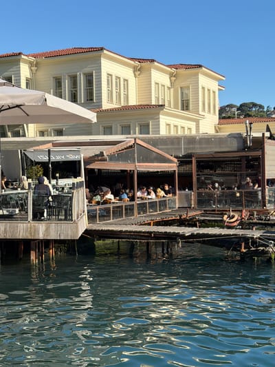 A waterfront scene in Üsküdar, Turkey, features a large, traditional wooden building with a red-tiled roof situated behind a contemporary, open-air restaurant complex built on stilts over the water. The restaurant has a glass-enclosed section and an open dining area with numerous tables and chairs occupied by patrons. In the foreground, the dark, rippling water reflects the sunlight, creating an abstract pattern of light and shadow. The sky is a clear, bright blue. The restaurant appears to be a popular dining spot, with many people seen eating and socializing at tables. A white umbrella is positioned to provide shade for one of the outdoor seating areas. The wooden structure of the restaurant extends out over the water, with a wooden walkway connecting different sections.