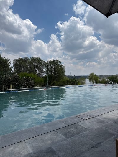 The image shows a view of a swimming pool located in Pueblo Nuevo de Jasso, Mexico, bordered by stone tiles on one side and a line of trees and cacti on the other. A lane divider floats in the water. The sky is partly cloudy with blue patches. In the distance, hills and a transmission tower are visible. The perspective is slightly tilted. A gray structure appears in the upper left corner of the frame.