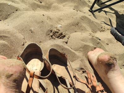 The image depicts a close-up view of sandy ground, likely a beach in Kandıra, Turkey, on a sunny day. Two pairs of sandals are visible. One pair of brown leather, open-toed sandals with straps is lying on the sand, partially covered in sand. The other sandal is a dark gray, possibly rubber, sandal, with its sole facing upwards. A human foot, partially covered in sand, is positioned near the brown sandals, suggesting someone was wearing them. The sand is coarse and appears dry, with some small debris scattered on its surface. In the background, the shadow of a dark, metallic object, possibly a chair or umbrella stand, is cast on the sand. The overall scene suggests a casual, outdoor setting, likely a beach environment.