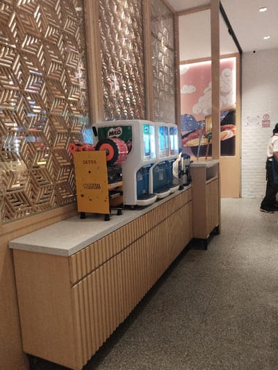 The image shows a beverage dispensing machine labeled "MILO" in a restaurant in Telukjambe, Indonesia, located at coordinates -6.301 latitude and 107.278 longitude. The machine is on a counter with a wooden cabinet and a white top surface, also on the counter is a smaller yellow machine labeled "CETRA".  The restaurant has wooden walls covered in a geometric pattern. The cabinet is made of light wood with a grooved vertical pattern, and the floor is covered in speckled gray tiles.  A second, larger, rectangular wooden cabinet is behind the dispensing machine, and a partially visible door with a wall covered in white tiles is on the right side of the image. There is a person in the background on the right side of the image, but the person is only partially visible.  The restaurant is clean, with a bright and modern aesthetic.
