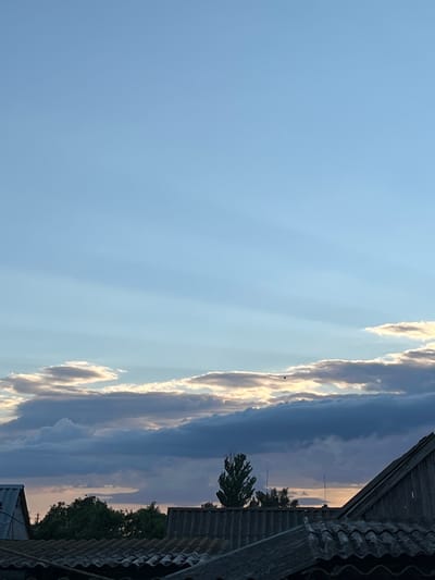 The image captures a twilight sky over Samiilove, Ukraine. In the foreground, the corrugated roofs of several buildings are visible, appearing weathered and dark. Above the rooftops, a line of lush green trees forms a dense canopy. Further up, a distinct, solitary tree with a pointed top stands out against the horizon. The sky itself is a canvas of pale blue, transitioning into streaks of lighter hues, possibly from the setting sun. Several cloud formations are present, some thin and wispy, others thicker and darker, obscuring parts of the sky. One small, dark object, possibly a bird or a balloon, is visible in the upper portion of the frame against a patch of bright clouds. The overall impression is one of a quiet, rural evening scene.