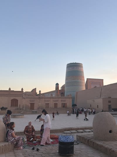 In the city of Khiva, Uzbekistan, within the Ichan Kala complex, a courtyard scene unfolds at dusk. In the foreground, a group of people are gathered on a rug laid out on the ground. A woman in a white shirt and pink pants stands while interacting with a younger woman sitting beside an older woman dressed in a patterned outfit. Two young girls in matching patterned outfits are also seated on a raised platform. A bottle of water and bowls are visible on the rug.

In the midground, a flat, paved courtyard is populated with other individuals. Several people are seen walking, and two children are riding bicycles. A dark-colored dog is also present in the courtyard.

The background is dominated by traditional Uzbek architecture. To the right, the distinctive Kalta Minor minaret, a cylindrical tower adorned with blue and white geometric patterns, stands prominently. Adjacent to it is a building with intricate tilework. Further to the left, other historical brick buildings with arched windows and doorways are visible, along with a minaret topped with a turquoise dome. The sky above is clear and a pale blue, indicating the late hour of the day. The overall impression is one of a historical site bustling with local life and tourists.