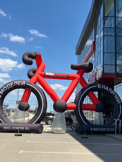 An inflatable red and black bicycle sculpture stands in front of a modern glass-fronted building under a partly cloudy blue sky. The sculpture is massive, with the wheels and frame dominating the foreground. Text is visible on the wheels, with "BIKE-CENTRE.RU" on the left wheel and "В НАЛИЧИИ" (meaning "in stock") and a phone number "8 800 500 03 20" on the right wheel. A red banner with "BIKE CENTER" and Russian text is also visible on the sculpture's frame. The building behind the sculpture appears to be a commercial establishment, with a prominent red and white sign visible on its facade. In the background, there is a busy highway with traffic. The scene is likely set in a retail or commercial area in Yablonovskiy, Russia, given the Russian text and the overall environment. There are no people clearly visible interacting with the sculpture.