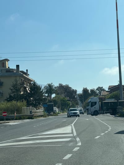 The image shows a street in Cisterna di Latina, Italy. The view is looking along the road, with the road surface taking up the right half of the frame. There are white lane markings and zebra crossing lines.
A white truck and a white car are visible on the road.
On the left side of the road, there is a building and trees. A blue road sign with "ROMA" and "VELLETRI" is visible. A red and white "Do Not Enter" sign is at the bottom left corner of the frame.
Overhead, there are power lines and a barrier arm with red and white stripes.
The sky is blue with some clouds visible.