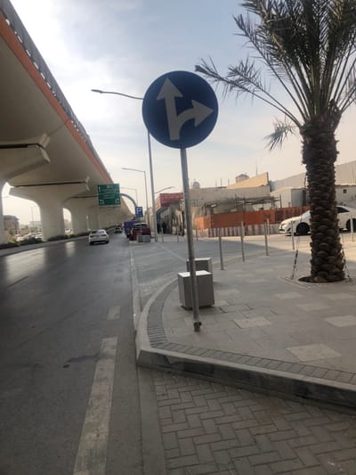 The scene depicts an urban street in Riyadh, Saudi Arabia, under bright, hazy daylight. The view is from a sidewalk, looking down a multi-lane asphalt road that recedes into the distance.
On the right side of the foreground, a paved sidewalk features alternating grey rectangular and herringbone-patterned pavers. A tall palm tree with a thick, textured brown trunk and green fronds stands rooted in the sidewalk, its base secured by a metal grate. A blue circular traffic sign, depicting white arrows for straight and right-turn options, is centrally mounted on a metal pole. Several grey metal bollards are spaced along the curb, which delineates the sidewalk from a vehicle area. Two grey cuboid structures are present on the sidewalk near the traffic sign pole.
On the left, a prominent elevated concrete highway structure, characterized by large, tapering light-grey support pillars and orange side barriers, extends into the background. Beneath this structure, a green overhead road sign is visible, displaying "Madinah Road" and "King Fahd Road" in both Arabic and English.
The asphalt road, appearing partially wet, shows visible white lane markings. A white sedan is observed traveling away from the camera in the left lane. A red pickup truck and other vehicles are parked or stationary on the right side of the road, adjacent to the sidewalk.
In the mid-to-far background on the right, a series of low-rise buildings line the street. Some buildings exhibit signs of construction or renovation, with visible frameworks and orange safety barriers. A tall, slender architectural feature, consistent with a minaret, is discernible in the distant background against the hazy sky. Indistinct dark figures, possibly pedestrians, are visible on the far right sidewalk in the mid-background.