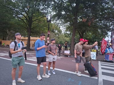 The image depicts an outdoor urban scene during daylight hours, consistent with a street bordering Lafayette Square Park in Washington, D.C., United States.
Six male individuals are prominently visible in the foreground and mid-ground. From left to right:
1. A male in a grey backward baseball cap, striped polo shirt, green shorts, and white sneakers stands on the asphalt, holding a black remote-like device and a white object. A black backpack is worn, and blue sunglasses hang from his shirt.
2. A male in a blue long-sleeve shirt, dark shorts, and white sneakers stands on the asphalt, looking down at a smartphone in his hands.
3. A male in a patterned t-shirt, light shorts, and a dark backward baseball cap, wearing white sneakers, stands on the asphalt, looking towards the right.
4. A male in a red "TRUMP" baseball cap, olive-green long-sleeve shirt, grey shorts, and light sneakers, with a brown sling bag, stands on the asphalt adjacent to a crosswalk.
5. A male in a red "TRUMP" baseball cap and a brown long-sleeve shirt stands near the crosswalk, extending both arms forward to hold a smartphone horizontally, appearing to record or take a selfie. He is simultaneously pulling a black, wheeled speaker unit featuring illuminated red, blue, and green lights on its side.
The environment includes an asphalt street with white crosswalk markings on the right and dashed white lines on the left. A reddish-brown paved area serves as a sidewalk or plaza. Numerous mature trees with green foliage form a dense canopy. A multi-lamp street light is visible. In the background, a light-colored building with tan walls is partially visible, along with parked green and white shared bicycles. On the far right, near the crosswalk, a vendor stall displays multiple flags, including several red "TRUMP" flags (some stating "TRUMP 2020"), a red flag with white text, and an American flag. Other merchandise is also visible on the stall. Several indistinct figures and small wagons are discernible in the deeper background.