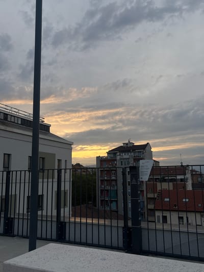 The image depicts a cityscape at dusk, possibly in Turin, Italy. Buildings of varying heights and architectural styles dominate the urban landscape. The sky is characterized by elongated clouds, tinged with the golden light of the setting sun. A metal railing or fence is visible in the foreground, partially obstructing the view. A gray pole is also visible on the left of the image. The buildings are a mix of residential and commercial structures, featuring brick, concrete, and painted facades. The presence of a ReLife sign on one of the buildings is an indicator of location. The lighting suggests a late afternoon or early evening scene, with a soft, ambient glow illuminating the surroundings.