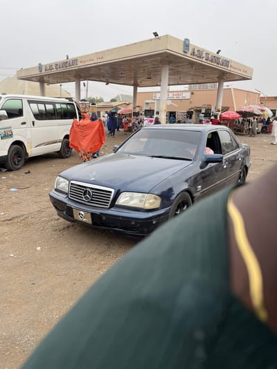 The image captures an active market scene in Kasuwan Kaji, Nigeria, adjacent to a gas station under an overcast sky.
In the immediate foreground, the right shoulder and a portion of the chest of an individual are visible, clad in dark green fabric with a vertical gold stripe.
A dark blue Mercedes-Benz W202 C-Class sedan occupies the central mid-ground, positioned at an angle facing the left. The vehicle's front grille features the Mercedes-Benz emblem, and a blank license plate bracket is affixed to the front bumper. The sedan is fitted with black multi-spoke wheels. A person is faintly visible in the driver's seat.
Behind the Mercedes, a white Toyota Hiace commuter van is present, oriented to the left. The van displays "0210" along with green and blue graphics on its side and also features black wheels.
Walking between the Mercedes sedan and the white van is a person draped in a bright orange garment, carrying multiple containers stacked on their head.
The background is dominated by an "A.M. KANKANI Nigeria Ltd." gas station, identifiable by its large canopy and prominent signage. Adjacent to the gas station, a building displays a sign for "A. R. MARKET," which also includes "BAUCHI STATE" in its text. Numerous market stalls, many topped with colorful umbrellas, are set up around this building. A multitude of individuals are observed moving and congregating throughout the market area.
The ground surface throughout the visible area is unpaved, appearing dusty and uneven.