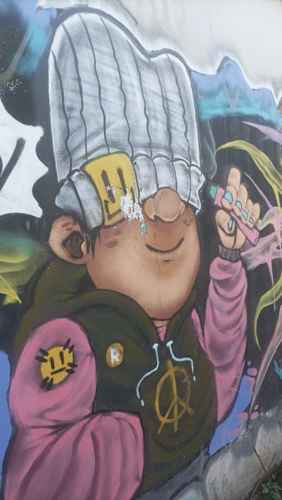 The image displays a close-up of a vibrant, multi-colored graffiti mural spray-painted onto a dark-toned wall, likely in an outdoor urban setting. The central subject is a stylized, cartoonish character with light brown skin, a prominent round nose, and eyes obscured by a large, vertically striped light grey and white knit beanie. A peeling yellow patch, featuring a rudimentary yellow smiley-like symbol, is affixed to the beanie, positioned above the character's right eye. The character wears a dark olive green hoodie, which is adorned with a large yellow peace symbol on the chest and a white stylized outline graphic. The character's left arm (viewer's right) is encased in a pink sleeve, upon which a yellow circular patch with a frowning smiley face is depicted. The right hand (viewer's left) is raised and holds a pink object with a light blue tip, resembling a marker or spray paint cap, with light blue fingernails visible on the fingers. The wall surface shows signs of wear, including a distinct vertical crack running through the character's face and horizontal cracks across the beanie. Areas of paint are peeling, notably on the yellow beanie patch. The background around the character consists of abstract splashes and outlines in various colors, including blues, yellows, and pinks. The lower right portion of the image reveals the base of a grey concrete structure and minimal green foliage, indicating a ground-level perspective. The specific location within the Special Capital Region of Jakarta, Indonesia, cannot be determined from the visual information provided.