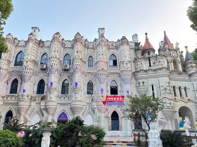 The image depicts a large, ornate building resembling a castle. The building is primarily white, constructed with what appears to be light-colored brick, and features numerous arched windows and decorative spires. Air conditioning units are visible on the exterior. There are several statues situated in front of the building, some white and classical in style, along with some trees and shrubs. The steps leading to the building are decorated with multicolored tiles. A sign with Chinese characters hangs above the entrance.

The location is identified as being in Baqiao, China.
