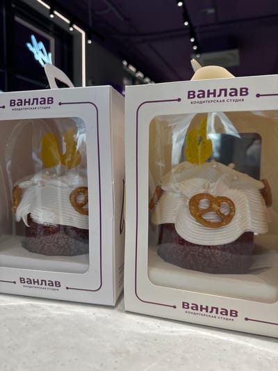 Two dessert items are packaged in individual white boxes with clear plastic windows. The boxes are labeled in Russian, with the words "Ванлав" and "КОНДИТЕРСКАЯ СТУДИЯ" visible, indicating a confectionery studio, presumably the brand of the dessert. The items appear to be decorated pastries or cupcakes, each adorned with white frosting, a pretzel, and yellow accents, possibly edible decorations. The pastries are placed on a surface, likely a store display or countertop, and are stacked one above the other. The pastries are in Kuleshovka, Russia.