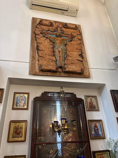 This image shows a wall in what appears to be a religious building, possibly a church or chapel, located in Sestroretsk, Russia.
Dominating the upper portion of the image is a large, relief carving on wood depicting the crucifixion of Jesus Christ. The carving features Jesus on the cross, with scrolled parchment-like textures surrounding him, inscribed with what appear to be religious symbols or script. A portion of an air conditioning unit is visible on the white wall above the carving.
Below this central piece, several framed religious icons are displayed on the wall. To the left, there are two icons, the upper one showing the Virgin Mary holding the infant Jesus, and the lower one depicting a similar scene with multiple figures. To the right, two more icons are visible. The upper right icon also features the Virgin Mary and child, while the lower right icon shows a woman in prayer or adoration.
In the center of the lower half of the image, a large, ornate wooden cabinet with glass doors is present. Inside, a hanging religious lamp (likely an icon lamp or censer) with blue glass elements is visible. The cabinet contains other religious artifacts, including what appears to be a large icon or painting behind the lamp. A string of pearls or rosary beads is also visible within the cabinet.
To the right of the cabinet, a vase with white flowers, possibly daisies, is placed on a ledge. Another framed icon is partially visible below the flowers.
The environment is dimly lit, with the light source appearing to come from above and from within the cabinet. The walls are white, and the overall impression is one of a sacred space adorned with religious art.