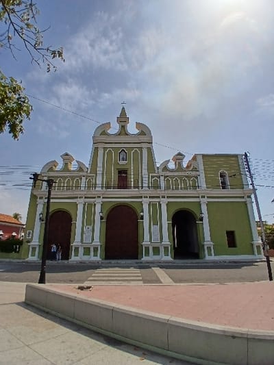 The image displays an exterior daytime view of a prominent two-story building located in San Joaquín, Venezuela. The structure features a light green facade accented by white architectural elements including pilasters, cornices, and balustrades. The central upper section is capped with a cross and contains a small statue within a niche, positioned above a red-brown door and balcony. The ground level exhibits three large arched entrances; the left two are equipped with dark brown wooden doors, while the rightmost is an open, dark passage. Two individuals, one in a blue top and one in a white top, are observed standing near the leftmost wooden door. In the immediate foreground, a paved asphalt street with a marked pedestrian crossing separates the building from a curved, raised concrete median with a reddish-brown top. A black streetlamp stands on the left side of the street. Overhead, electrical wires are visible against a partly cloudy blue sky, with tree branches occupying the upper left corner of the frame.