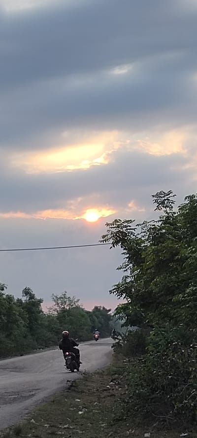 The image depicts a rural road scene in Blangmajron, Indonesia, at dusk. The sky is filled with dramatic clouds, with the sun partially visible through them, casting a warm glow. On the paved, somewhat unkempt road, a person is riding a motorcycle, their form silhouetted against the light. Another motorcycle is visible further down the road, also moving away from the viewer. The roadside is lined with lush green trees and foliage, creating a natural, somewhat overgrown environment. A power line stretches horizontally across the upper portion of the scene, adding a man-made element to the otherwise natural landscape. The overall impression is one of a peaceful, end-of-day journey through the Indonesian countryside.