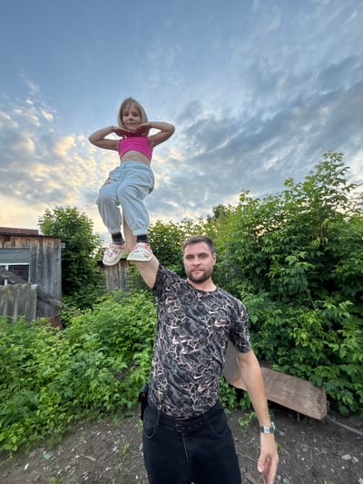 In an outdoor scene set in Маркидоновка, Russia, a man and a young girl are captured in a dynamic pose against a backdrop of lush greenery and a cloudy sky. The man, wearing a black patterned t-shirt and dark pants, extends his right arm, his hand pointing forward. He wears a watch on his left wrist. The girl, positioned in front of him, is balancing on what appears to be a wooden fence or railing. She is wearing a pink sports bra, light-colored pants, and distinctive white clogs with colorful designs. Her arms are raised above her head, hands near her face.

The low angle of the shot emphasizes the sky, which is filled with scattered clouds, suggesting it is either early morning or late afternoon. The surrounding environment is characterized by dense green foliage and hints of wooden structures, indicative of a rural or suburban setting. The overall mood appears playful and energetic, with the two subjects engaged in an activity that showcases balance and perhaps trust. There is no visible text in the image.