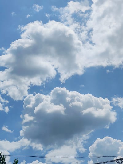 The image depicts a bright, clear sky filled with fluffy white cumulus clouds. The sky is a vibrant blue, suggesting it is daytime. The clouds are varied in size and shape, with some appearing dense and voluminous, while others are wispy and scattered. There are no people or prominent objects visible in the sky itself, but at the very bottom of the frame, a thin black wire stretches horizontally, and some dark green leaves from a tree are visible on the left. This suggests the photo was taken from ground level, looking upwards. The overall impression is one of a pleasant, sunny day with fair weather. The image conveys a sense of openness and vastness associated with the sky. There is no visible text in the image.