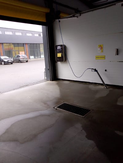 This is an image of the interior of a car wash facility, likely located in Saransk, Russia. In the foreground, a wet concrete floor with a drainage grate is visible. To the right, a metal stand holds a car wash wand and hose. The wall behind it has several signs, including one with a cartoon pig and the text "Не буль..." and another yellow sign with the text "ВОДА+ХИМИЯ". On the left side of the image, the open entrance to the car wash reveals two cars parked outside, with a building featuring large windows and a sign that reads "АРЕНДА". The overall environment appears to be an indoor, functional space for vehicle cleaning, with evidence of recent use due to the wet floor.