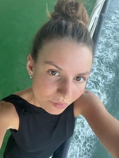 A close-up selfie captures a young woman with her hair in a messy bun, her brown eyes looking directly at the camera. She is wearing a black sleeveless top, and her right shoulder and arm are visible. She has multiple small earrings in her left ear. The background is a mix of green water on the left and turbulent blue-green water with white foam on the right, suggesting she is on a boat or ferry. A dark railing or edge of the vessel runs diagonally through the right side of the image, with white supports visible behind it. The scene appears to be outdoors, likely on the water. The image does not provide specific details to pinpoint the exact location within Batas Desa Skotong Timur, Indonesia.