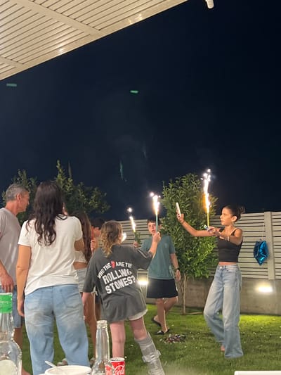 A group of people stand outside at night, holding sparklers. A woman in a black top and jeans is in the background next to a white fence. There is a blue balloon by the fence. Another person has a walking cast on their right leg and is wearing a Rolling Stones t-shirt. There are several plastic bottles on a table in front of the group. Two green lights appear in the dark sky.
