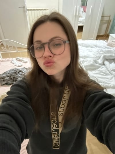 A young Caucasian female with long brown hair and round, dark-rimmed glasses is depicted from the chest up, taking a selfie indoors in what appears to be a bedroom located in Scafati, Italy. She is wearing a dark-colored hooded sweatshirt. A beige lanyard with "Calvin Klein jeans" printed repeatedly in dark brown text is visible around her neck, hanging over the sweatshirt. Her lips are pursed in a "kissing" or "pouting" expression.

The background reveals a domestic setting. To the left, a white metal headboard of a bed is partially visible, with a light pink sheet and some rumpled clothing (including a plaid shirt) and white socks on the bed. To the right, a significant portion of an unmade bed covered with white bedding, appearing rumpled, occupies the lower right quadrant. Behind the bed, a white wardrobe with multiple doors, one featuring a mirrored surface reflecting the bed, is present. A white radiator is visible on the wall to the left, behind the subject's head. The room has light-colored walls and flooring.