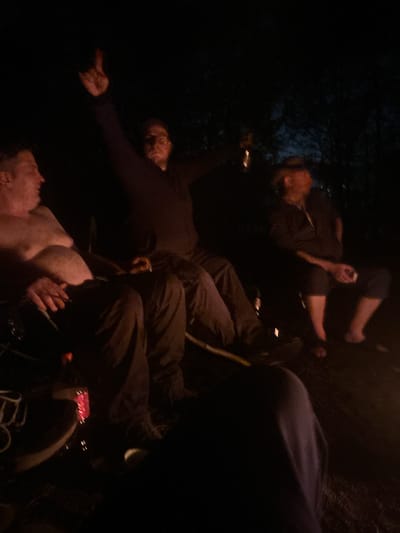 The image appears to depict a group of individuals in a poorly lit outdoor setting at night. There is a mix of people, some partially obscured. One man is shirtless, lying on the ground, while others are standing or positioned around him. Clothing colors are dark, suggesting a night-time setting. The environment seems to be natural, possibly a park or wooded area. The image doesn't provide enough context to ascertain the precise location within Юзуфоўскі сельскі Савет, Belarus.
