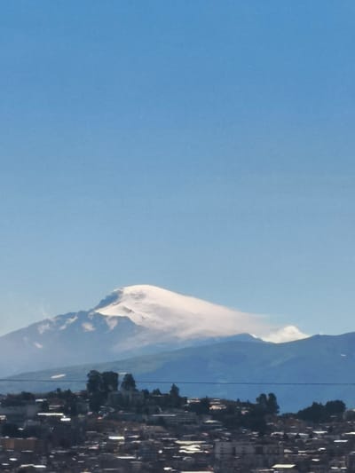 The image captures a scene from the city of Quito, Ecuador, with a distant view of a snow-capped mountain against a clear blue sky. The foreground features an urban landscape composed of closely packed buildings. A cluster of trees and possibly a church structure is seen in the middle ground between the urban area and the mountain. Powerlines stretch across the view at mid-level. The mountain is partially covered by clouds.