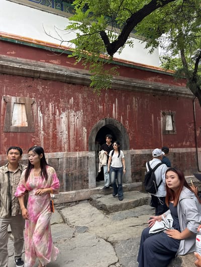 The image depicts a bustling scene at a historical site within the Haidian District of China, likely the Summer Palace (Yiheyuan), characterized by traditional Chinese architecture. A prominent, weathered red wall with peeling paint and vertical streaking forms the backdrop, featuring an arched entrance near its center. Two recessed, rectangular, off-white panels, resembling sealed windows, are embedded in the red wall, one to the left and one to the right of the archway. Above the red wall, a horizontal stone band supports a tiled roof section with green and orange tiles, topped by a white wall and a more ornate, multi-colored roof structure (partially visible) featuring blue and green patterns.
Numerous individuals of East Asian descent are present. In the immediate foreground, a man in a light-colored striped shirt and beige pants walks alongside a woman wearing a flowing, long pink and white tie-dye patterned dress. She holds an orange device, possibly a smartphone, attached to a strap. Both are moving from left to right.
Through the arched entrance, a man in a dark t-shirt and light pants is visible, followed by a woman in a white t-shirt, blue jeans, and white-framed sunglasses. Further into the archway, other indistinct figures can be seen. To the right of the archway, several people are navigating irregular stone steps. One man, seen from behind, wears a white baseball cap, a light striped shirt, and a black backpack.
In the foreground right, a woman with reddish-brown hair is seated on a stone step, looking towards the right side of the frame. She wears a light grey zip-up jacket over a dark blue or grey jumpsuit or wide-leg pants and holds a partially visible clear plastic water bottle. The ground consists of uneven stone paving and steps. Large, verdant trees with dense foliage are visible in the upper right and upper left portions of the frame, providing shade and partially obscuring the upper parts of the building and sky. A thick tree trunk is prominent in the upper right.