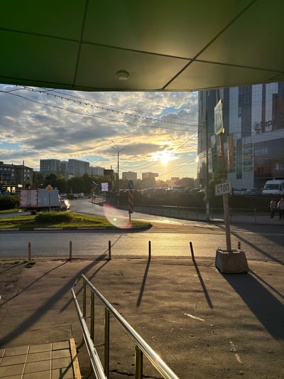The image captures a street scene in Pushkino, Russia, during what appears to be late afternoon or early evening, given the low angle of the sun and long shadows. The sky is filled with dramatic clouds, illuminated by the setting sun, which creates a warm, golden light across the asphalt road.

In the foreground, a building with a dark green facade is on the left, with geometric paneling. To the right, a metal railing curves away from the viewer, alongside a tiled surface. The road itself is wet in places, reflecting the light and suggesting recent rain or ongoing street cleaning. Several metal bollards are scattered along the roadside, and a concrete block with a metal fixture sits near the edge of the road.

A street sign with the Russian word "СТОП" (STOP) is prominently displayed on a pole, indicating a traffic stop. A pedestrian crossing sign is also visible nearby. In the middle ground, cars are present on the road, suggesting traffic. Further in the distance, a cityscape unfolds with numerous apartment buildings of varying heights, characteristic of urban development. The overall atmosphere is one of a typical urban environment transitioning from day to evening, with a visually striking natural light display in the sky.