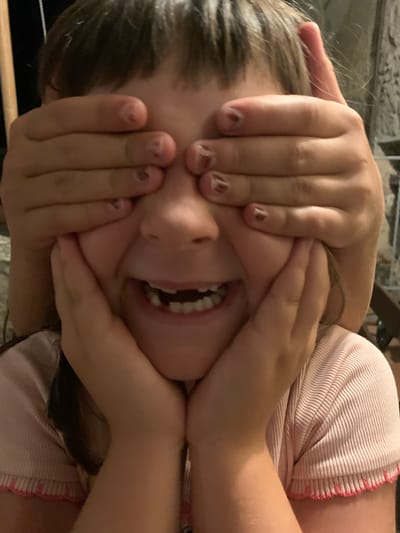 A young girl, her eyes covered by two sets of hands, smiles widely, revealing missing front teeth. The hands, likely belonging to two different people, are positioned in front of her face. She wears a pink ribbed shirt with a scalloped hem. The background is dark and out of focus, suggesting an indoor or evening setting. The image appears to be a candid shot of a playful moment.