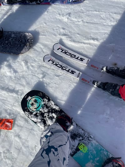 The image displays a close-up, ground-level view of a snow-covered outdoor area in Melchsee-Frutt, Switzerland, illuminated by daylight. In the immediate foreground, a person's lower leg, wearing white snow pants with a grey and blue abstract pattern, is positioned with a red-accented boot secured into a black and teal snowboard. The snowboard's top surface is partially covered with fresh snow, revealing a circular teal-rimmed sticker depicting a smiley face with palm tree eyes. To the right of the snowboarder's leg, a pair of white Stöckli Axis skis, marked "SWISS MADE" and "149," lie parallel on the snow, with the bindings and parts of black ski boots, one with a red cuff, visible. Further left and partially obscured, the tail of another snowboard, featuring a grey and black pattern resembling snakeskin, is visible on the snow. In the background, at the top of the frame, the tip of a blue and purple ski with faint white text is also present. The snow surface is uneven, showing tracks and disturbances consistent with an active winter sports setting.