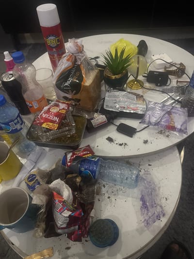 The image shows a cluttered white circular table with various items scattered across its surface. In the foreground, a blue mug is visible, next to crumpled wrappers and debris. A plastic bottle of water lies on its side, and further back, another blue mug and a packet of milk are present.
On the upper level of the table, a red and white can of BNC Mosquito Spray stands upright. Next to it, a loaf of bread in plastic packaging is visible, along with packets of Nescafe. A small artificial plant in a gold-trimmed pot is also on this level. Several plastic bottles, including a Fanta bottle, are scattered around. Other objects include a watch, a power adapter, and various food wrappers and debris.
The overall impression is one of disarray, with remnants of food and drink items and general clutter. The image does not contain enough information to determine a specific location within Dutsen Alhaji, Nigeria, or to infer any actions or interactions beyond the presence of the scattered objects.