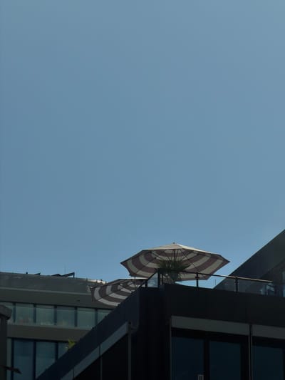 This media file is a low-angle shot of a building and the sky. The building appears to be modern, with dark glass windows and sleek metallic accents. The sky is a clear, pale blue, suggesting it is daytime.
Prominently featured are two striped umbrellas, one red and white, the other white with brown stripes, attached to the exterior of the building. One umbrella is fully visible, while the other is partially obscured. The umbrellas are angled as if to provide shade on an outdoor balcony or terrace. A hint of greenery, possibly palm fronds, is visible peeking out from behind one of the umbrellas, suggesting a warm climate or a well-maintained urban space.
The perspective looking up towards the building and sky creates a sense of height and openness. The lighting suggests it is daytime, possibly mid-morning or afternoon, given the clear sky and moderate lighting. There are no people visible in the frame. The scene evokes a sense of urban calm and architectural interest. There is a small white sticker with black text on the window of the building, but the text is illegible. The location context provided is Frankfurt, Germany.