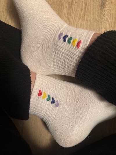 The image displays a close-up, top-down view of two feet, each wearing a white ankle sock, resting on a light brown wooden floor composed of planks. Both socks feature a repeated embroidered pattern on the outer ankle region, consisting of five small, arc-shaped elements. The colors of these elements, from left to right on the top sock, are purple, blue, green, yellow, and red. The bottom sock shows the same pattern, oriented such that the colors appear in reverse order from left to right (red, yellow, green, blue, purple) when viewed from the perspective of the foot's inner side. The socks exhibit minor pilling consistent with use. Portions of dark, ribbed fabric, presumably pants or clothing, are visible around the ankles and outer edges of the feet. The lighting suggests an indoor setting. The location in the city of Visp, Switzerland, cannot be determined from the visual evidence provided in the image.