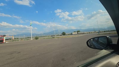 The image depicts a wide, paved lot with a mountainous backdrop under a bright blue sky with scattered white clouds. In the foreground, a car's side mirror is visible on the right, reflecting the surroundings. The lot appears to be a rest stop or parking area, with a few parked vehicles including a red truck on the left and several other trucks and cars on a road in the distance. A tall pole with a green and blue logo stands prominently in the middle ground, along with streetlights and road signs. The landscape includes green fields and sparse trees leading up to rolling mountains. The overall scene suggests a rural or roadside location in the Жамбыл ауылдық округі, Kazakhstan.
