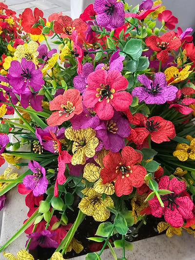 The image displays a close-up, overhead view of an arrangement of vibrant artificial flowers, predominantly in shades of red, pink, yellow, and purple, with green foliage. The flowers have multiple petals, many with a textured pattern, and dark centers. The arrangement is densely packed, with some flowers overlapping others. The background is blurred, but suggests a light-colored, possibly textured surface, and hints of a dark potting medium at the bottom. The scene appears to be an indoor or sheltered outdoor setting, likely a decorative element within a home or establishment. No specific location in Laval, Canada can be identified from the visual information provided.