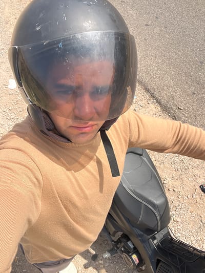 The image is a top-down shot of a person sitting on a motorcycle. The person is wearing a black helmet with a clear visor and a beige sweater. The motorcycle is black. The ground is a mix of asphalt and gravel. The location appears to be a roadside in an outdoor area. While the image does not provide explicit information to confirm the city of Beni Mellal, Morocco, based on the surrounding clues, one can suggest that the photo was taken there.