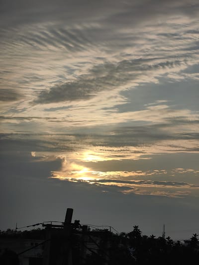 The image captures a dusky sky over Gharaunda, India, with the sun partially obscured by clouds. The clouds are layered, with some exhibiting a striated pattern. In the foreground, silhouetted against the sky, is a power pole with electrical wires and fixtures. A line of trees forms the horizon, with hints of buildings visible behind them. The overall lighting suggests either early morning or late evening.