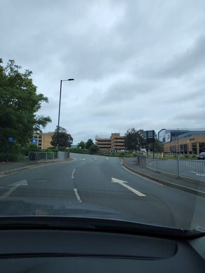 The image captures a ground-level view from inside a vehicle on a cloudy daytime in Peterborough, United Kingdom. The scene depicts a multi-lane asphalt road curving gently to the right, with a white arrow painted on the tarmac indicating a right turn. The vehicle's dashboard is visible at the bottom of the frame.
On the left side of the road, dense green trees and bushes line a pavement, partially obscuring buildings behind them. A tall, black street light with a modern LED fixture stands prominently. Visible signs include a blue circular sign with a white bicycle symbol, indicating a cycle route, and a blue rectangular warning sign featuring a white triangle with an exclamation mark and text that appears to read "BRIDGE" or "BRIDGE STREET," likely referring to the railway bridge or a bridge over the River Nene.
On the right side, a silver metal pedestrian barrier separates the road from a paved area and further vegetation. Beyond this, several large buildings are visible. These include a multi-story car park constructed of brown brick with visible concrete ramps, which corresponds to the Queensgate/Railway Station Car Park. Adjacent to this, a modern commercial building complex, identifiable as part of Rivergate Shopping Centre, features various materials including brickwork, glass, and grey cladding. Signage for "Greene King" and "VUE" (a cinema chain) is discernible on these buildings. A dark-colored car is partially visible on the far right lane, heading away from the camera. The sky is uniformly overcast with light grey clouds, indicating a dull weather condition. No people are visible in the frame.