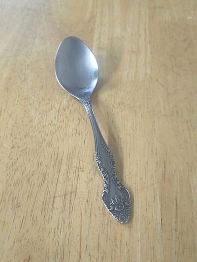 A single silver spoon lies diagonally on a light brown wooden surface. The spoon is positioned with the bowl at the top left of the image and the handle extending towards the bottom right. The bowl of the spoon is oval-shaped and reflects the ambient light. The handle is ornately decorated with floral patterns and embossed details. The wooden surface appears to be a tabletop or a similar flat wooden material with a visible wood grain pattern. There are no people, animals, or other objects present in the image. The lighting appears to be overhead, casting subtle shadows. The image is taken from a top-down perspective.