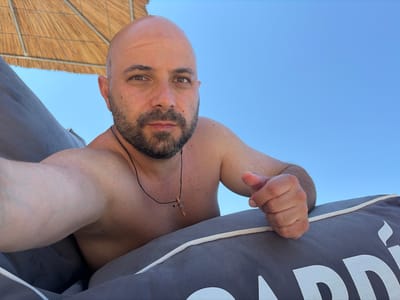 The image shows a bald, bearded man taking a selfie outdoors. He is wearing a necklace with a small wooden cross. The man is shirtless, and his skin appears wet, suggesting he may have been swimming. He is lying down on a gray cushion, and behind him, a thatched umbrella and a clear blue sky are visible. The scene appears to be a beach or a resort area. Based on the context provided in the prompt, the location is identified as the city of Kassandra Municipal Unit, Greece. The man is looking directly at the camera with a neutral expression. His right arm is bent, and his hand is resting on the cushion. The lighting suggests it is daytime, possibly midday, given the bright sunlight. The overall atmosphere is relaxed and summery.
