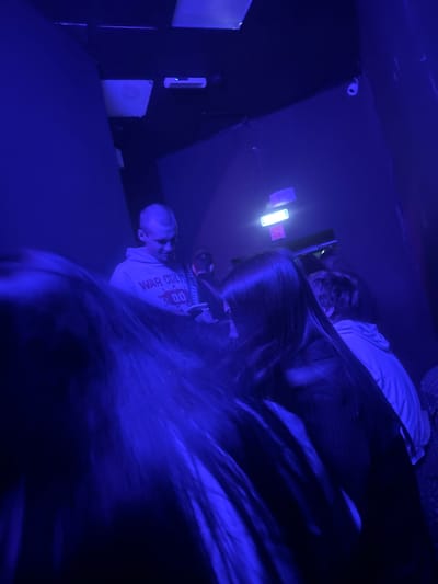A dimly lit indoor space, bathed in blue light, is captured with multiple people present. The focus is on a young man wearing a grey hooded sweatshirt with the words "WAR CULT" and the number "00" visible on the front. He is looking down at a smartphone he holds in his hand. Several other individuals are visible, some blurred due to motion, with long dark hair obscuring their faces. The environment appears to be a club or a similar entertainment venue, with overhead lighting fixtures visible. The overall impression is that of a candid shot taken during a social gathering.
