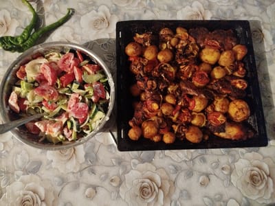 A table is laid with a floral tablecloth, set with two dishes of food. To the left, a silver bowl is filled with a chopped salad containing tomatoes, cucumbers, and onions, dressed in a creamy sauce. A fork rests in the salad. To the right, a black baking tray holds roasted meat and vegetables, including small potatoes, tomato slices, and onions. The food appears to be home-cooked. In the upper left corner, two wrinkled green peppers lie on the tablecloth. The scene suggests a meal is about to be served or has recently been enjoyed. The lighting indicates it is daytime. The setting is Ak-Suu, Kyrgyzstan. There is no visible text in the image.