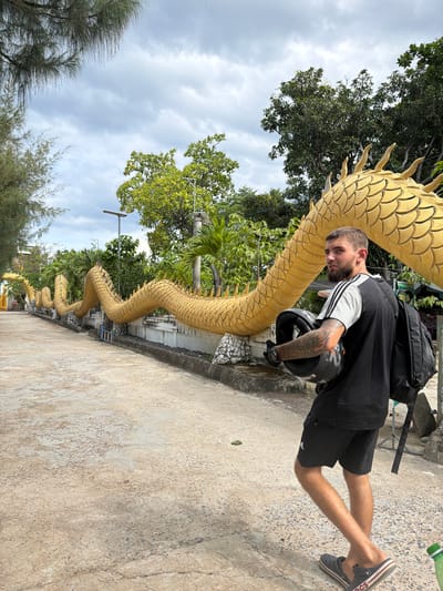 An outdoor, daytime shot in Cam Ranh, Vietnam, features a man standing on a concrete path next to a long, winding golden dragon statue. The image is taken from a slightly low angle, looking up towards the man and the dragon, with a cloudy sky and abundant green trees forming the background.
The man, with a beard and visible tattoos on his arms, is wearing a black t-shirt with grey and white striped sleeves, black shorts, and dark Crocs sandals. A black backpack is worn over his shoulders, and he holds a black motorcycle helmet with orange accents in his left hand. He is looking directly towards the camera with a neutral expression.
The prominent golden dragon statue snakes along the edge of the concrete path. Its body is covered in detailed scales, and it features a series of spines running down its back. In the distance, more segments of the dragon statue are visible, suggesting it extends for a significant length.
The surroundings include lush green trees with dense foliage, some palm trees, and what appears to be utility poles or light fixtures. The concrete path shows some wear and texture. A partially visible green bottle is seen near the man's feet in one of the crops. The text "CROCS" is visible on the strap of his sandal. The overall scene suggests a cultural or tourist attraction, likely a temple or garden, in Vietnam.