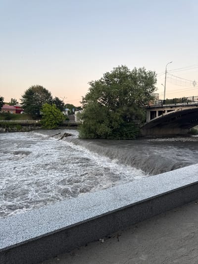 This image depicts a wide river flowing through an urban area, with a visible man-made waterfall or dam in the foreground. The water is turbulent and appears to be a grayish-brown color, indicating it may be carrying sediment. On the left bank of the river, a park-like area with lush green trees is visible, leading up to some buildings with red roofs. Further in the background, a bridge spans the river, supported by concrete pillars. The bridge has a railing with decorative elements, and overhead power lines are strung across. On the right bank, more trees and vegetation are present, with a concrete embankment in the foreground of the image. The sky is a pale, hazy gradient of light blue and pink, suggesting either dawn or dusk. There are no people clearly visible in the immediate foreground or midground, though a few small figures can be vaguely discerned on the riverbank on the left. The overall scene captures a natural element integrated into a developed urban environment. The location is identified as the city of Askhazan, Russia.