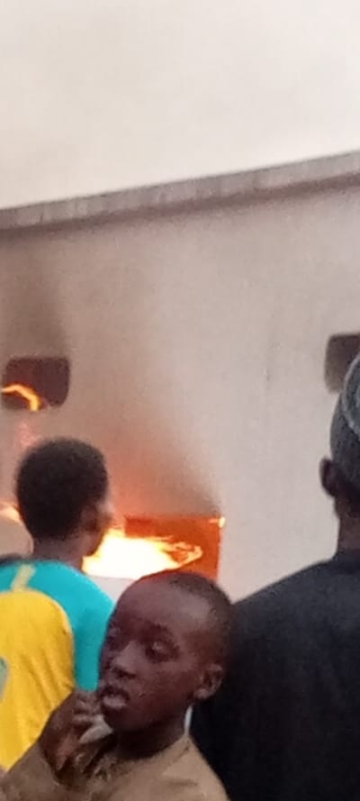 The image depicts an indoor scene featuring three individuals observing a source of intense heat or fire. In the foreground, slightly to the left, is a young boy with dark skin and short, dark hair, wearing a light brown top. He is partially turned towards the right, with his gaze directed towards the bright light in the background. His left hand is raised towards his mouth, suggesting he might be pondering, chewing, or making a gesture.
Behind the boy, to the left side of the frame, is another person, seen from behind. This individual has dark skin and short, dark hair and is wearing a bright blue top with a yellow section on the back or shoulder. They are also looking towards the background.
To the far right, partially visible, is the back of an adult's head and shoulder. This person is wearing a dark cap or hat and a dark garment, and is also facing towards the background.
The background is dominated by a large, light-colored wall or structure, possibly made of brick or concrete, which appears to be part of an industrial oven, furnace, or kiln. There are at least two openings in this structure that emit a very bright, orange-yellow light, indicating the presence of fire or extreme heat. One larger, rectangular opening is visible prominently in the mid-ground, with another smaller, higher opening to its left. The bright light from these openings casts a warm glow on the surroundings.
The overall impression is one of people observing a high-temperature process, possibly in a bakery, workshop, or industrial setting. The curious expression of the boy and the focused posture of the adults suggest an engagement with the activity involving the fire. No text is visible in the image.